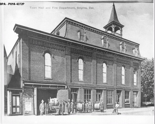 An image of the Smyrna Old Town Hall building, which previously held the library and Opera House. A two-story, old brick building with a spire at one side.